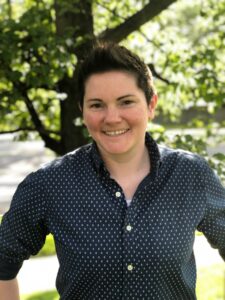 Picture of Dr. Kathryn Kein, a white woman with short brown hair wearing a dark blue button up shirt. The shirt has small white polkadots and white buttons. She is standing in the center of the frame smiling at the camera. There is a tree blurred out in the background